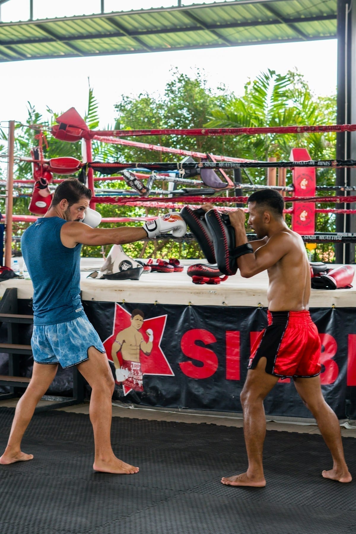Two athletes training Muay Thai techniques at an outdoor gym near a boxing ring in Thailand