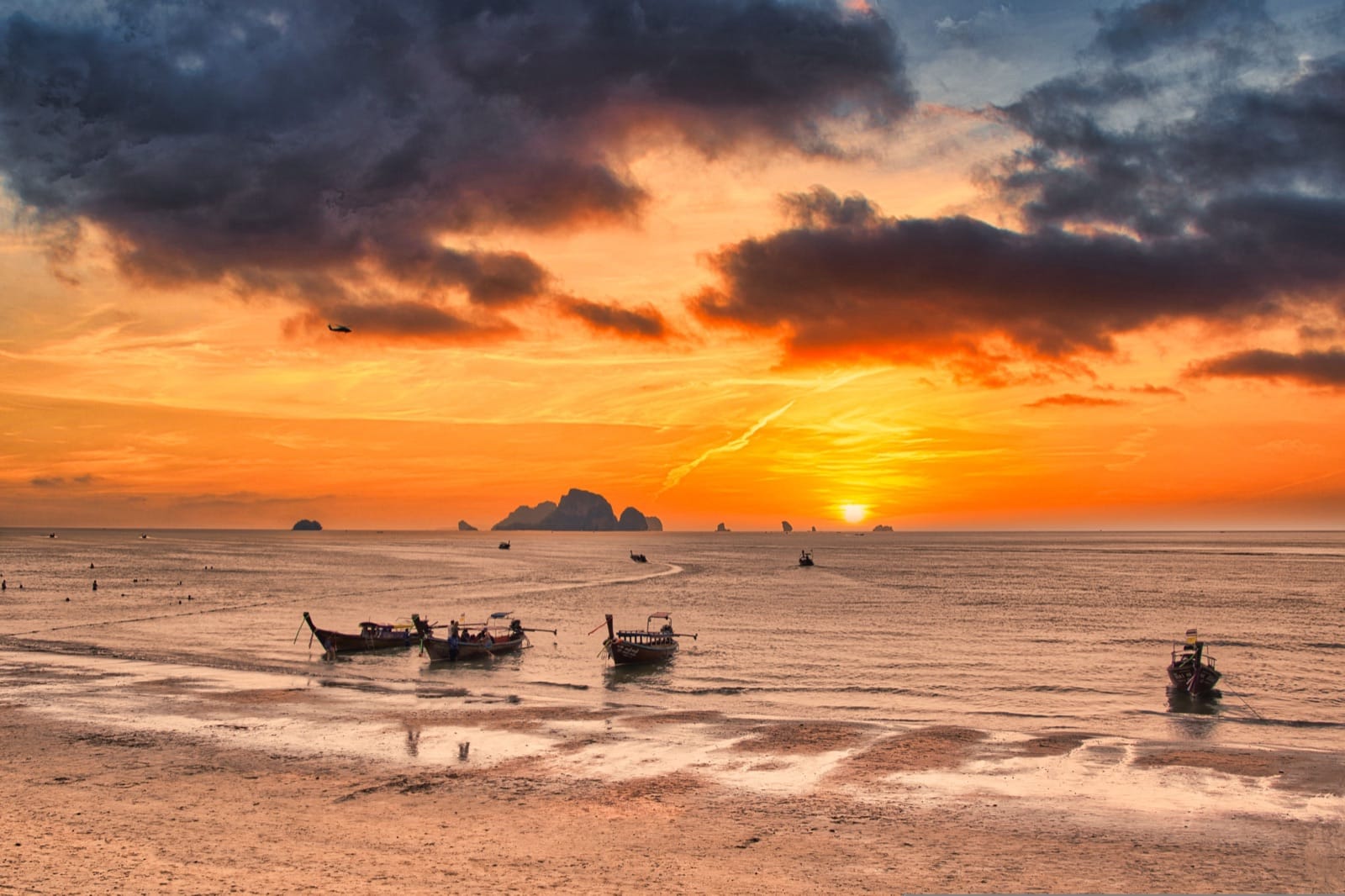 Dramatic sunset over a Krabi beach with longtail boats silhouetted against orange sky and limestone islands