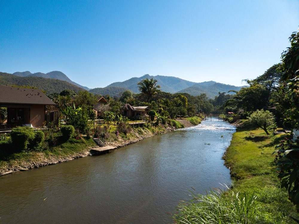 Fluss schlängelt sich an Gästehäusern und grünen Ufern vorbei, dahinter Berge in Nordthailand