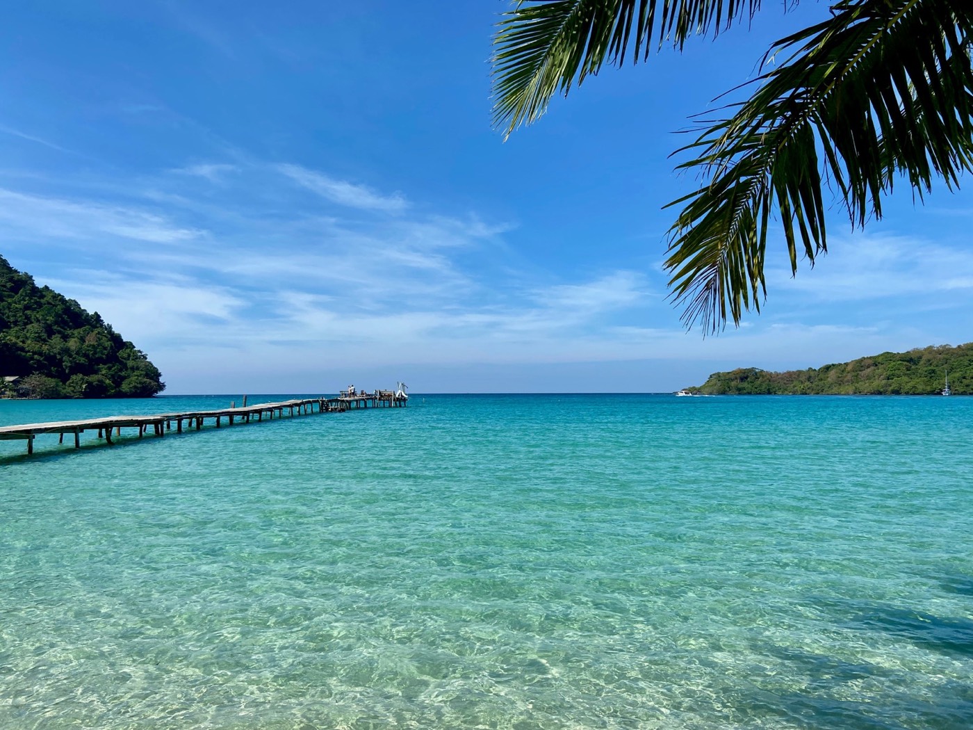 Long wooden pier extending over crystal clear turquoise water framed by palm fronds, Ko Kut, Trat, Thailand