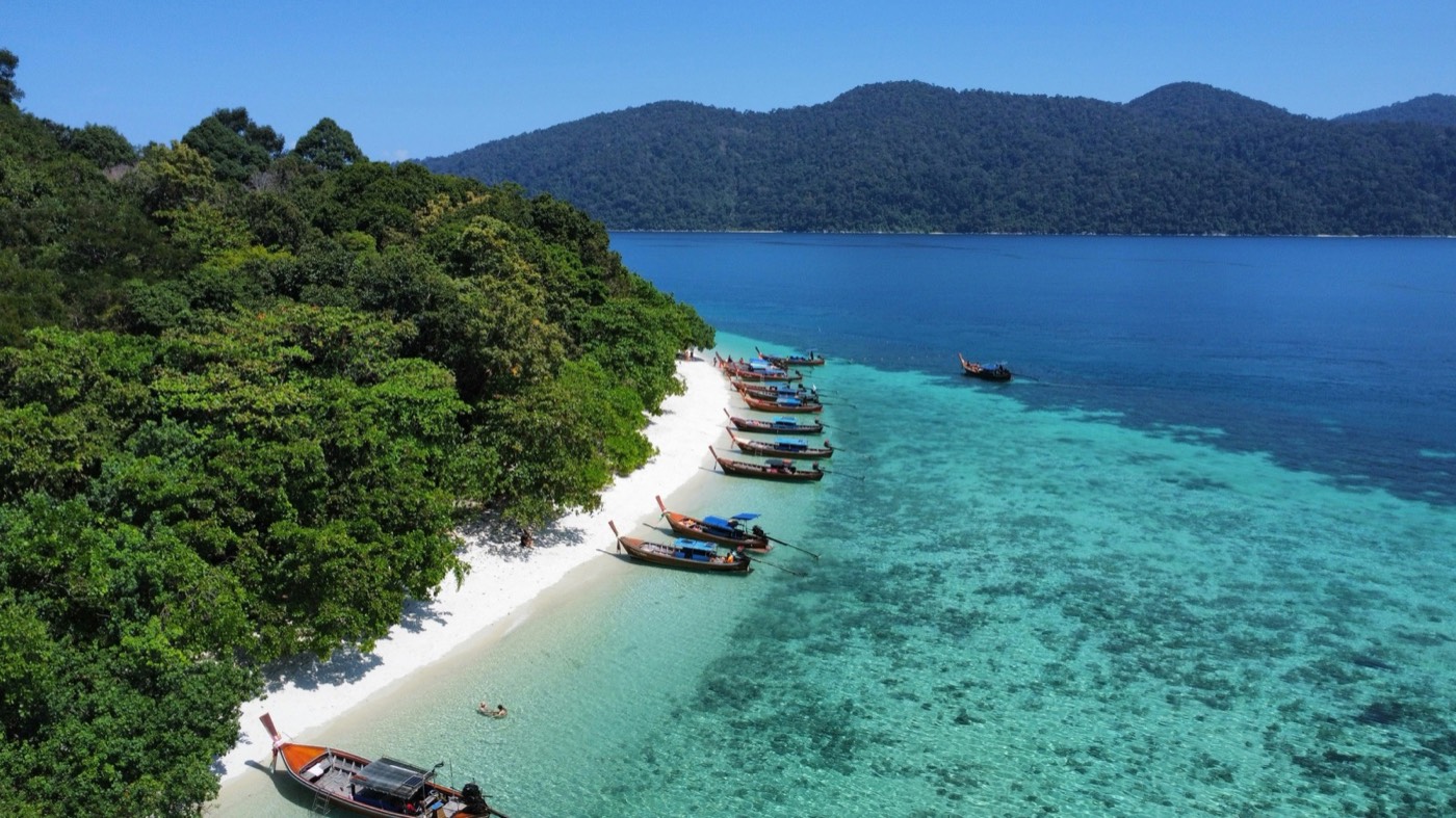 Longtail boats lined up on a white sand beach backed by lush green hills, Koh Lipe, Satun, Thailand